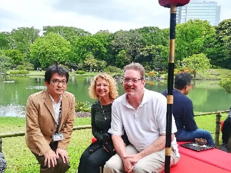 Three people sitting on a bench in a Japanese garden, with greenery and a pond in the background.