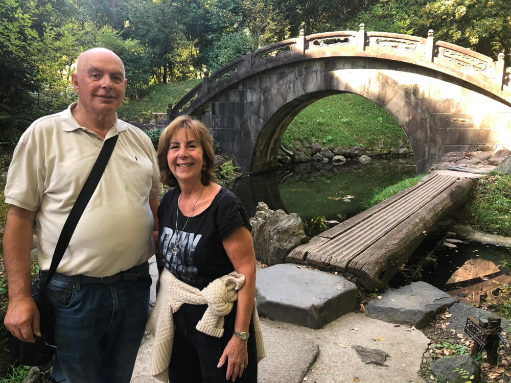 A couple standing together in front of an old stone bridge, surrounded by nature.