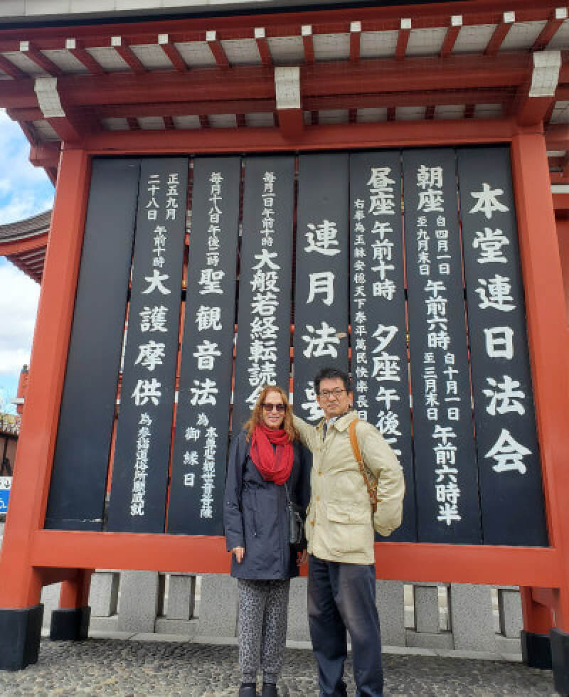 Two people standing in front of a traditional Japanese temple sign with large kanji characters.