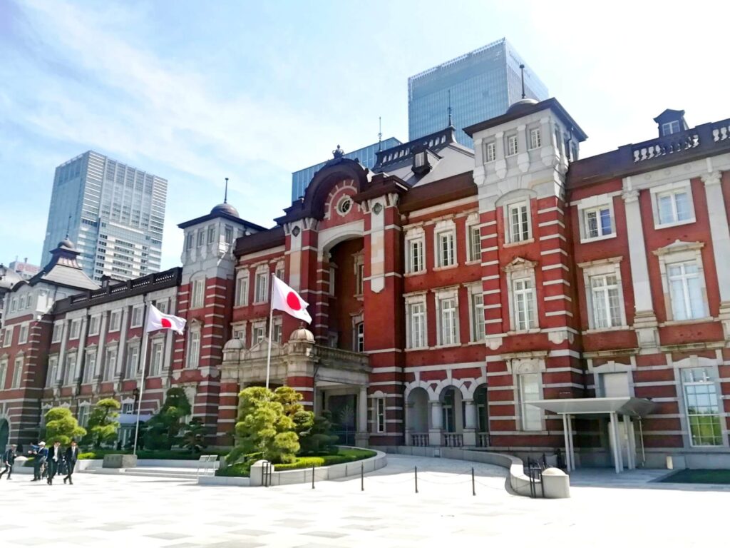 The historic red brick Tokyo Station building, with Japanese flags displayed outside, set against a backdrop of modern skyscrapers.